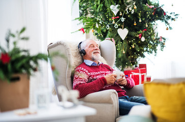 A portrait of senior man with headphones sitting on armchair at home at Christmas time.