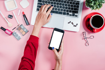 Woman hands holding blank screen smartphone and using laptop on pink background with laptop,cosemetic product and cup of coffee.Top view,Flat lay.