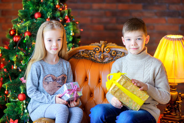 Fototapeta na wymiar brother and sister are sitting on the armchair and holding gifts, against the background of the celebratory Christmas tree