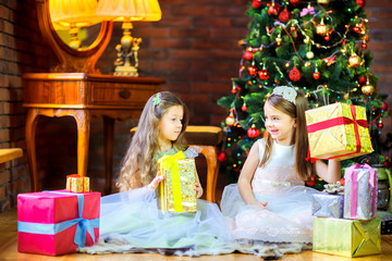 girls in beautiful dresses sitting on the floor near the Christmas tree give each other gifts
