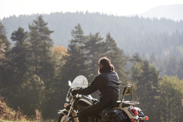 Back view of bearded biker with long hair in black leather jacket sitting on cruiser motorcycle on...