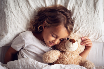 Portrait of a young girl (kid) sleeping in her bed and hugs a teddy bear.