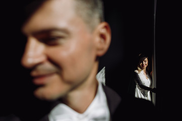 Handsome groom standing before a bright window while bride waits on the background