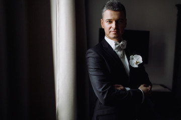 Groom's morning. Handsome man in black tuxedo with white orchid in his pocket stands before a...