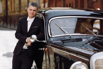 Handsome groom in black tuxedo poses before a black retro car somewhere in old town