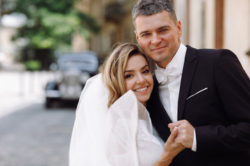 Stylish bride and groom look charming walking before a black retro car along the street