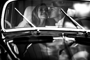 Fototapeta na wymiar Look from the outside at adorable wedding couple in classy dresses sitting inside a black retro car and hugging each other tender