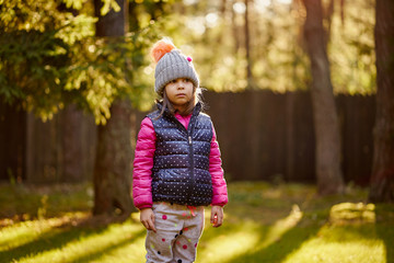 lovely little girl in a cap and jacket stands on the grass in a park in an autumn forest