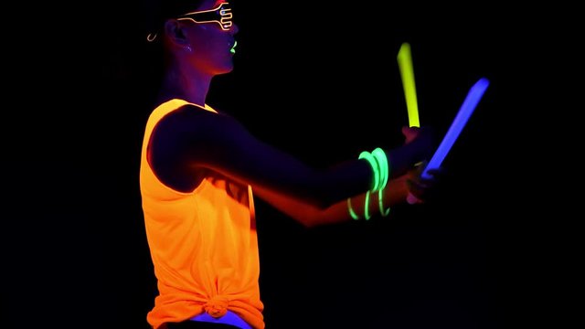 Beautiful sexy woman with UV face paint, glowing clothing, glowing glasses, bracelet dancing in front of camera holding chemical light, half body side shot. Asian woman. Party concept.