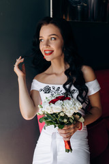 Lovely bride with dark hair and red lips holds white wedding bouquet in her arms sitting on a sofa