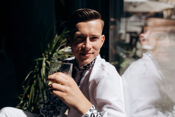 Handsome young man in white suit with embroidery sits on a windowsill before bright window