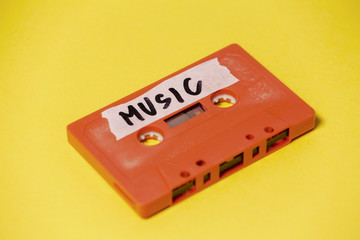 A vintage cassette tape (obsolete technology), orange on a yellow surface, angled shot, carrying a label with the handwritten text Music.
