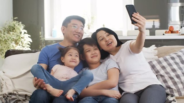 Handheld tracking shot of happy Asian family of four sitting on sofa and taking selfie