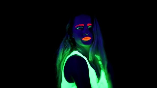 Beautiful sexy woman with UV face paint, glowing clothing, glowing bracelet dancing in front of camera. Caucasian woman. Party concept.