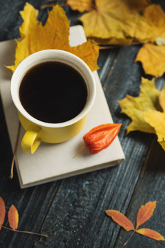Yellow coffee mug and book on a dark wooden table with autumn scarf and leaves
