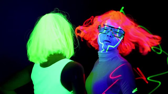 Beautiful sexy women with laser, UV face paint, wig, glowing glasses, glowing clothing dancing together fast in front of camera, Half body shot. Caucasian and asian woman. Party concept.