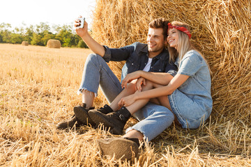 Naklejka na ściany i meble Photo of young couple man and woman smiling while sitting under big haystack in golden field, during sunny day