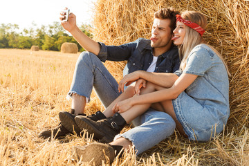 Naklejka na ściany i meble Photo of countryside couple man and woman smiling while sitting under big haystack in golden field, during sunny day