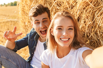 Photo of cheerful couple man and woman taking selfie while sitting under big haystack in golden field, during sunny day