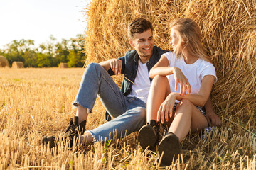 Naklejka na ściany i meble Photo of young couple man and woman sitting under big haystack in golden field, during sunny day