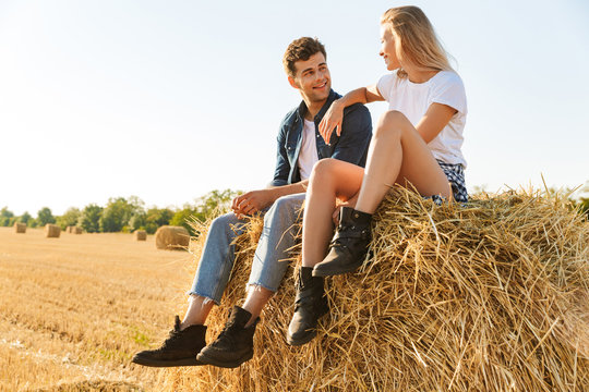 Photo of romantic couple man and woman sitting on big haystack in golden field, during sunny day