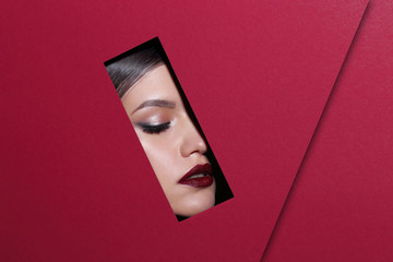 Face of young beautiful girl with a bright make-up and red lips looks through a hole in red paper.