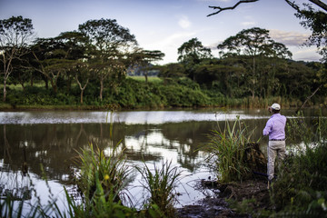 Landscape scene of farm dam with late afternoon reflections on the still water. Rear view of man assembling fly fishing rod. In the distance trees are dotted with weaver bird nests. Farmland beyond. 