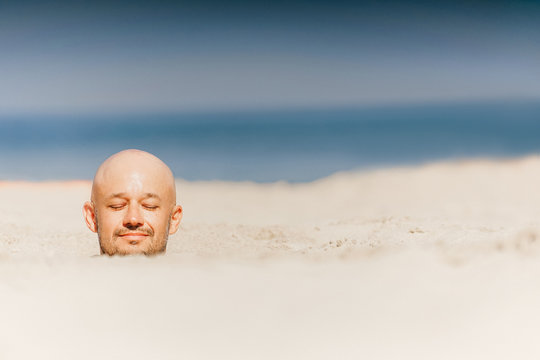 Male bald head above sand.  Man buried  alive in desert. Happy person relaxing in uninhabited beach on sun. Closeup portrait of funny guy taking sunbath with body under ground near ocean. Therapy.