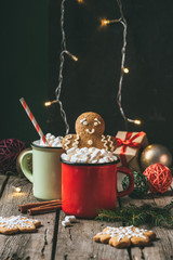 two cups of cocoa with marshmallows and gingerbread man on wooden table with christmas light garland