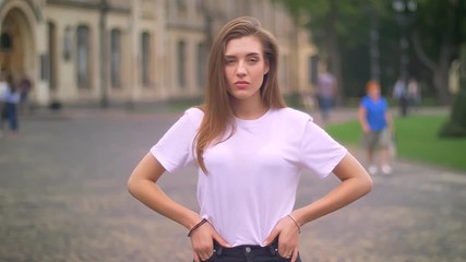 awesome caucasian female is standing with her hands on hips and assured look at camera, city background, daytime