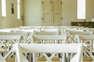 Partial view of white French cross-back timber chairs arranged for an event inside an old building. Seating is set up for Wedding Ceremony. No people. 
