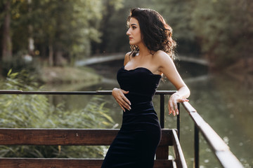 Stunning young woman in seductive long black dress with naked shoulders stands on the bridge over a lake in the rays of evening sun