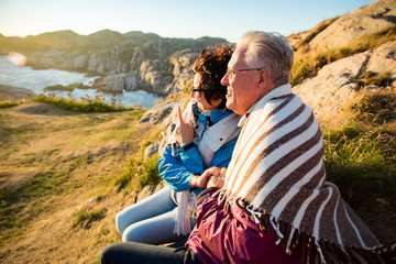 Loving mature couple hiking, sitting on windy top of rock, exploring. Active Mature man and woman wrapped in blanket, hugging and Happily smiling. Scenic view of sea, mountains. Norway, Lindesnes.