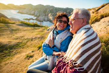 Loving mature couple hiking, sitting on windy top of rock, exploring. Active Mature man and woman wrapped in blanket, hugging and Happily smiling. Scenic view of sea, mountains. Norway, Lindesnes.