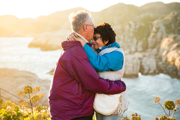 Loving mature couple traveling, standing on the top of rock, exploring. Active man and woman hugging and kissing, Happily smiling. Scenic view of mountains and sea on sunset. Norway, Lindesnes