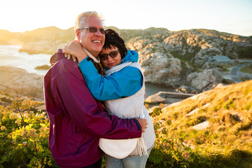 Loving mature couple traveling, standing on the top of rock, exploring. Active man and woman hugging and kissing, Happily smiling. Scenic view of mountains and sea on sunset. Norway, Lindesnes