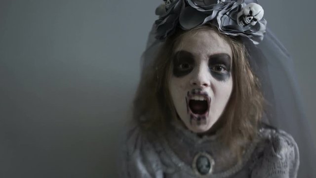 Dracula bride The girl in the costume of Halloween, in the makeup of the dead man screams and looks terribly at the camera. On the head of a child is a wreath of skulls and gray veil