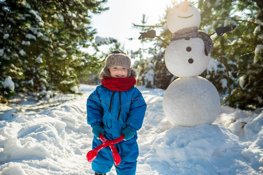 Little boy plays in snow and makes snowballs near large snowman