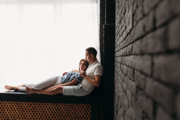 Young man and woman hug each other tender sitting on the windowsill in a bright room