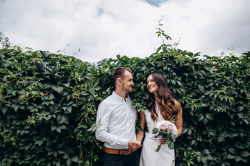 Tender hugs of wedding couple standing before an old brick wall covered with green ivy