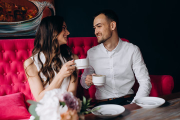Charming young wedding couple drinks coffee sitting on a bright pink couch in a cosy European cafe