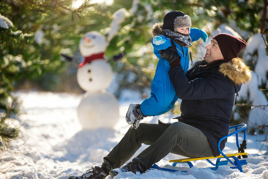 Father sits on sledges and holds little son in snow winter wood