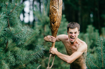 Odd primitive naked man with huge wooden stick hunting in forest. Adult male have fun like crazy psyche with rude club in hands. Expressive excited boy face.  Wild masculine strength. Cruel warrior.