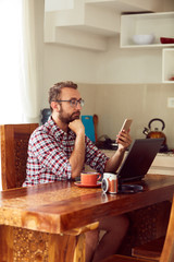 Modern bearded man using cellphone and drinking coffee with laptop.