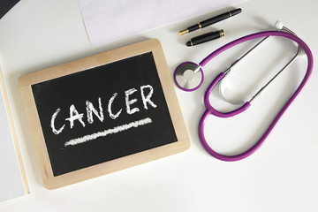 Word CANCER written on a blackboard next to a stethoscope and a pen on medical consultation desk.