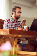 Modern bearded man using cellphone and drinking coffee with laptop.