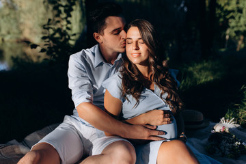 Charming young expecting couple rest on the plaid under green tree in the rays of evening sun
