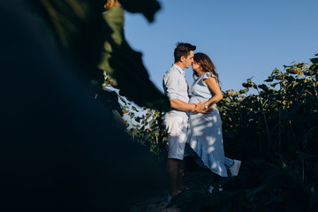 Cheerful man and pregnant woman hug each other tender standing in the field with tall sunflowers around them