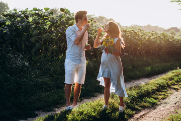 Expecting man and woman walk along the path across the field with sunflowers in beautiful summer evening