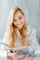 beautiful smiling young woman in pajamas reading book in bedroom